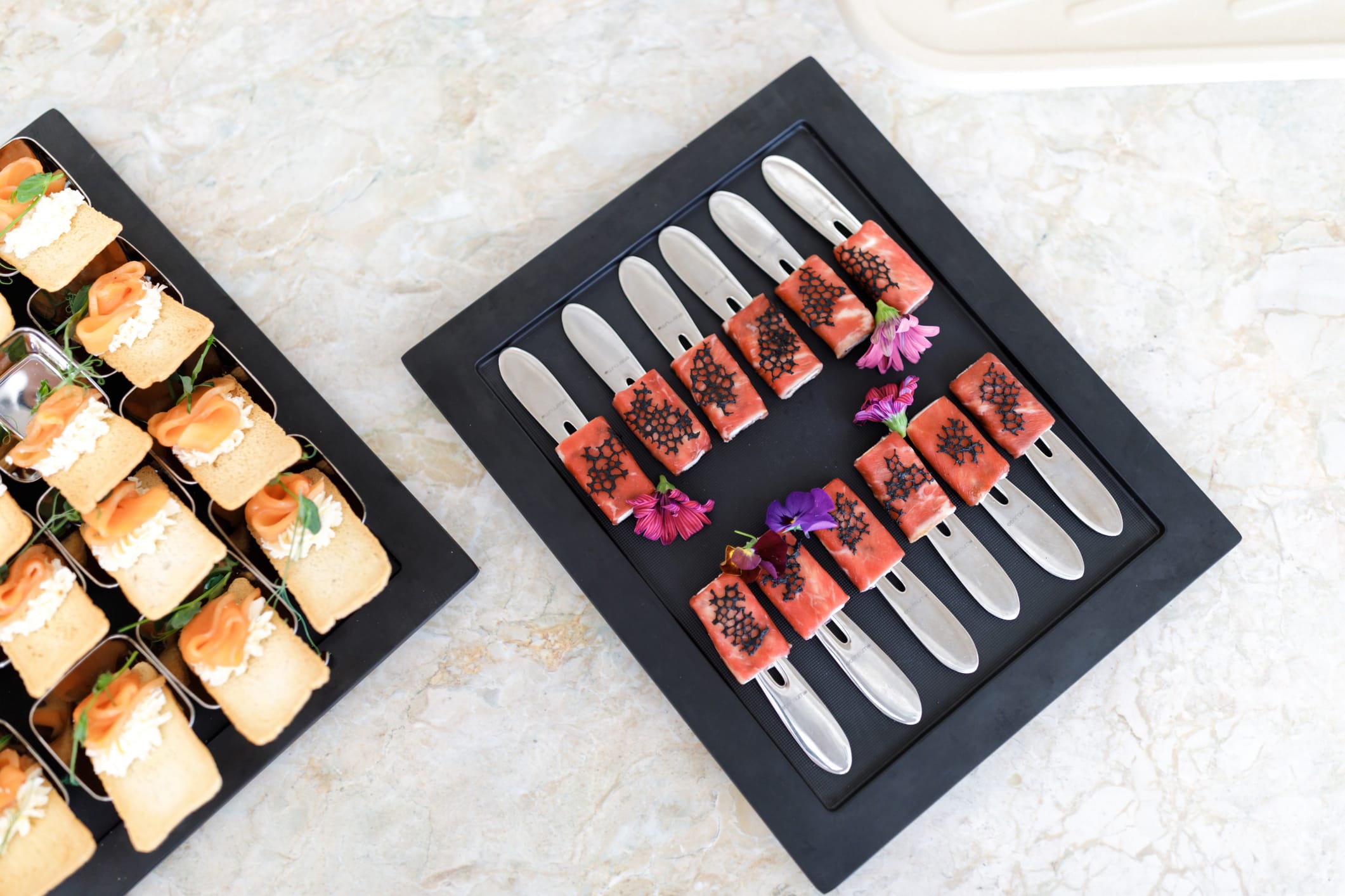 Two black trays of canapes, smoked salmon tartines and garnished bites with edible flowers, on a marble surface.
