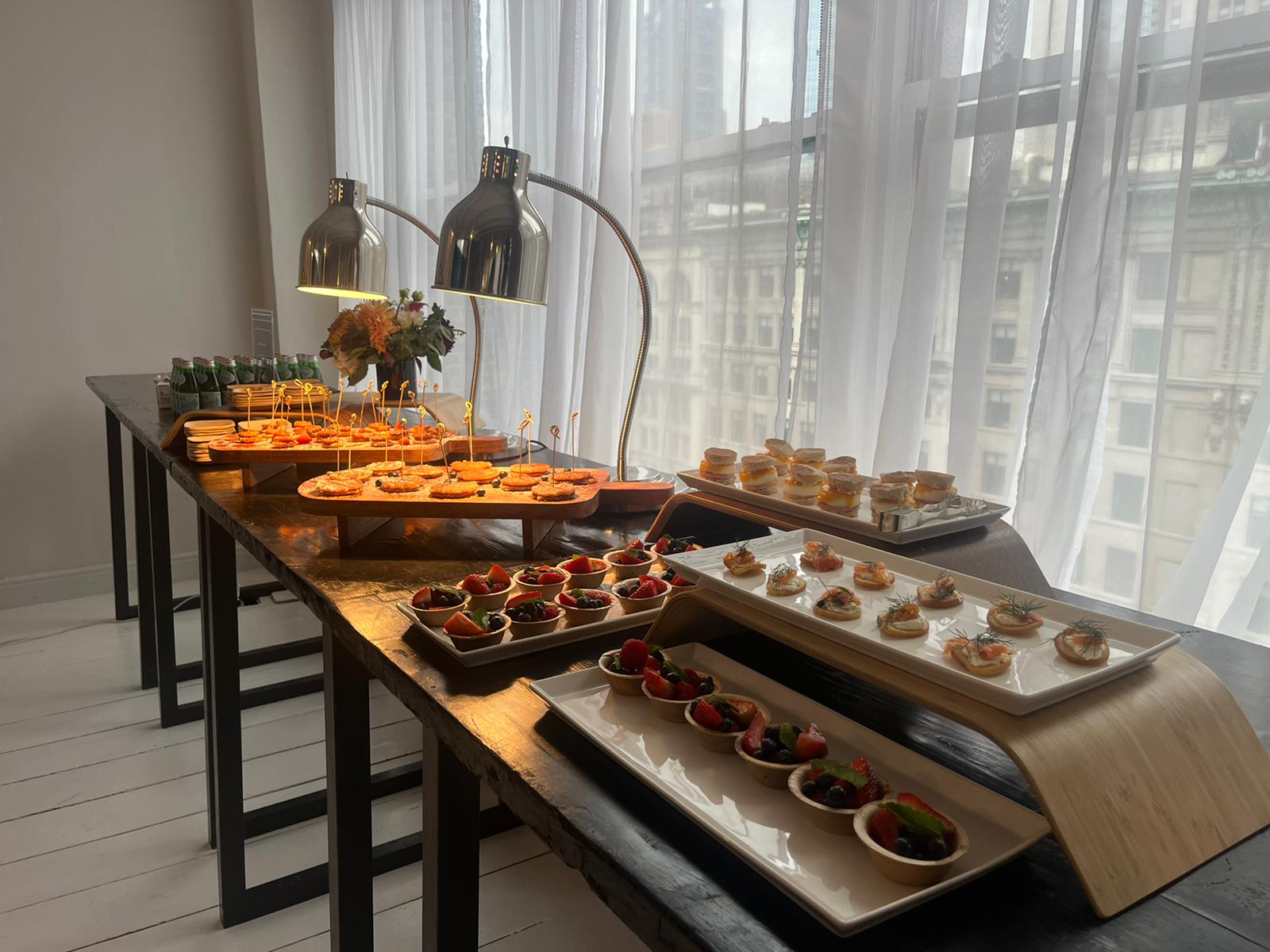 Daytime office spread under heat lamps with skewered bites and tartlets beside tall windows