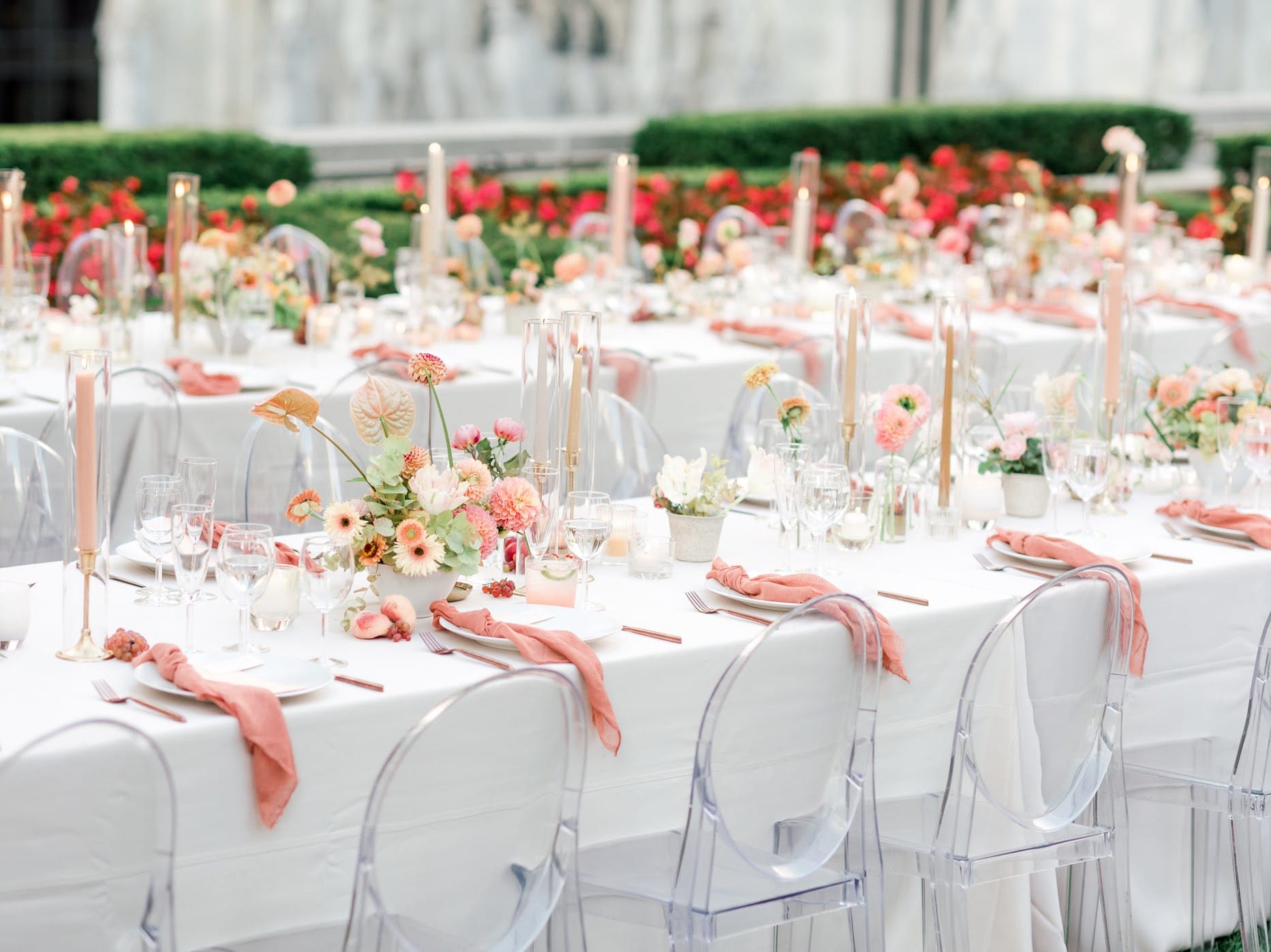 Outdoor wedding reception with long tables, ghost chairs, and coral floral centerpieces in a garden setting