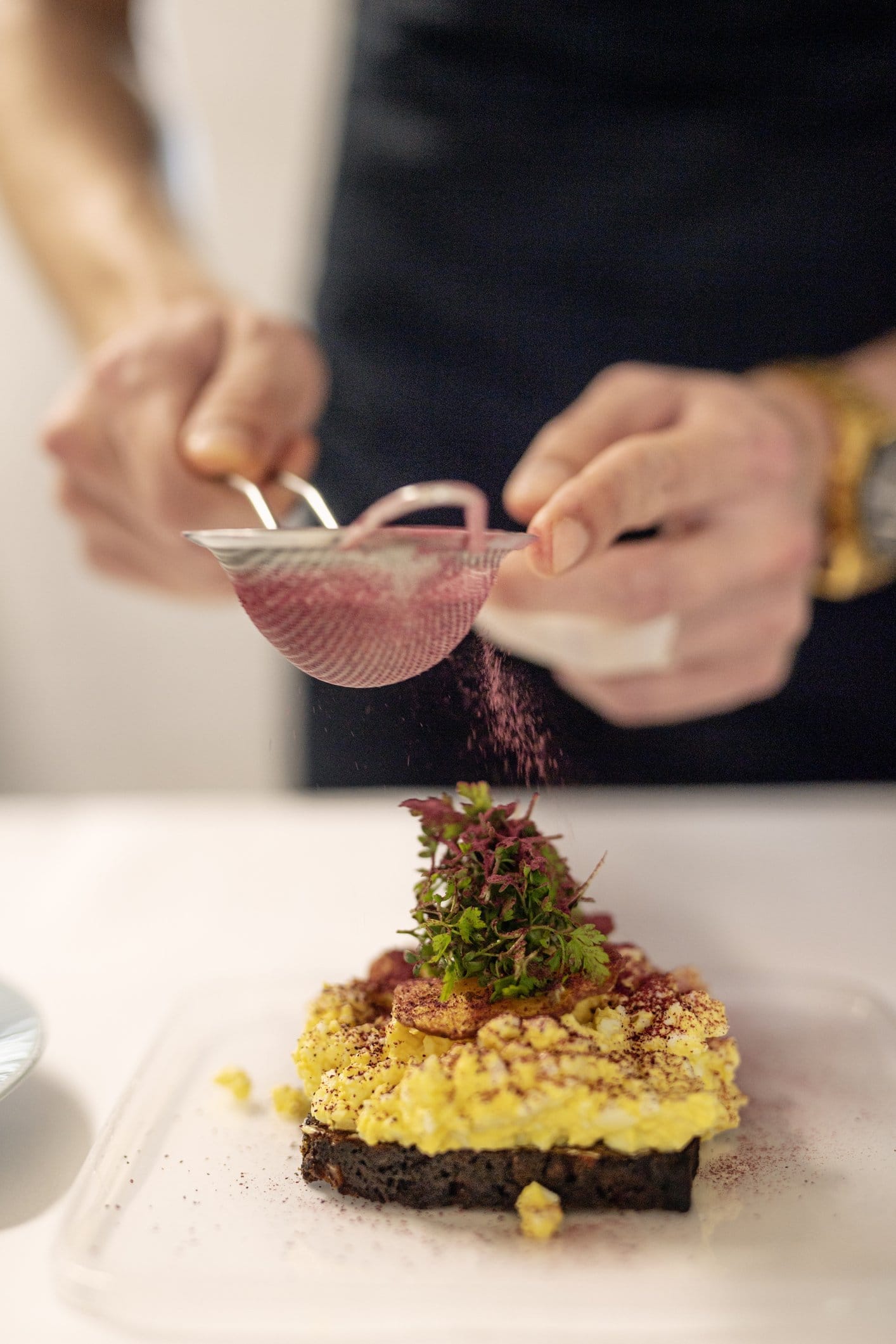 Chef dusting finishing powder through a sieve over a canapé on dark rye