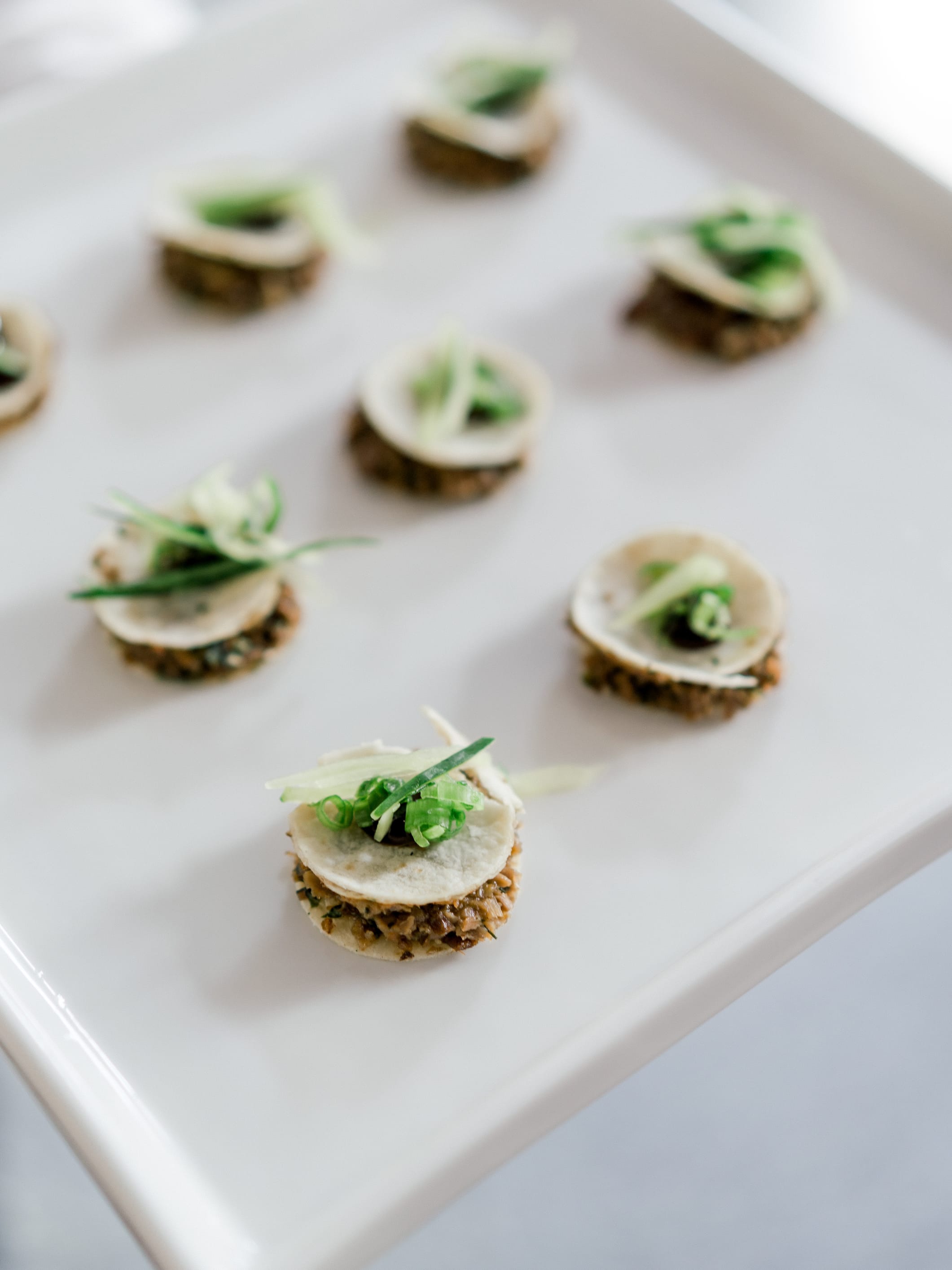 Mini pancake canapés topped with shaved cucumber and scallion, in rows on a white platter