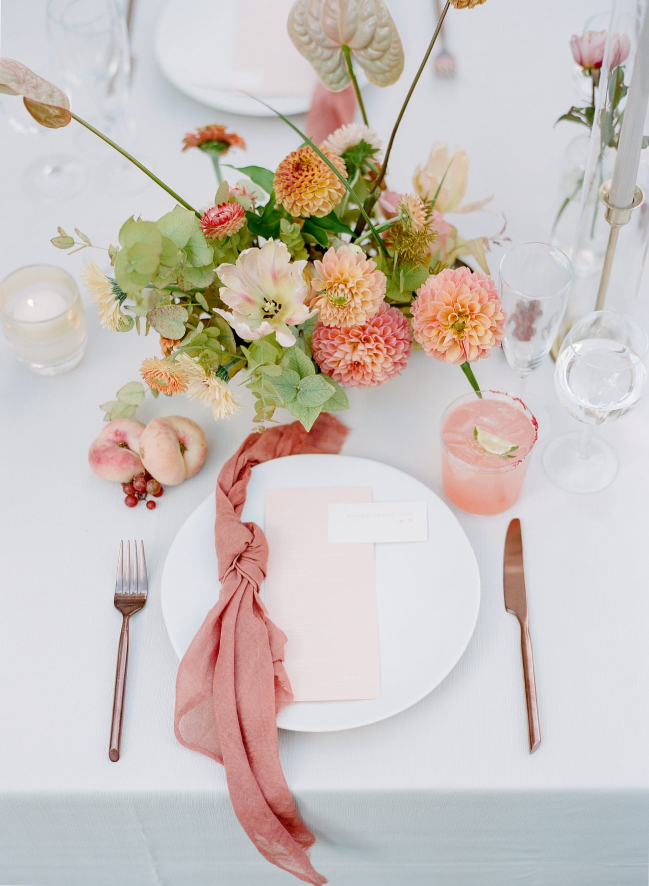 Overhead place setting with a knotted rose napkin, blush menu card, copper cutlery, and peach dahlias